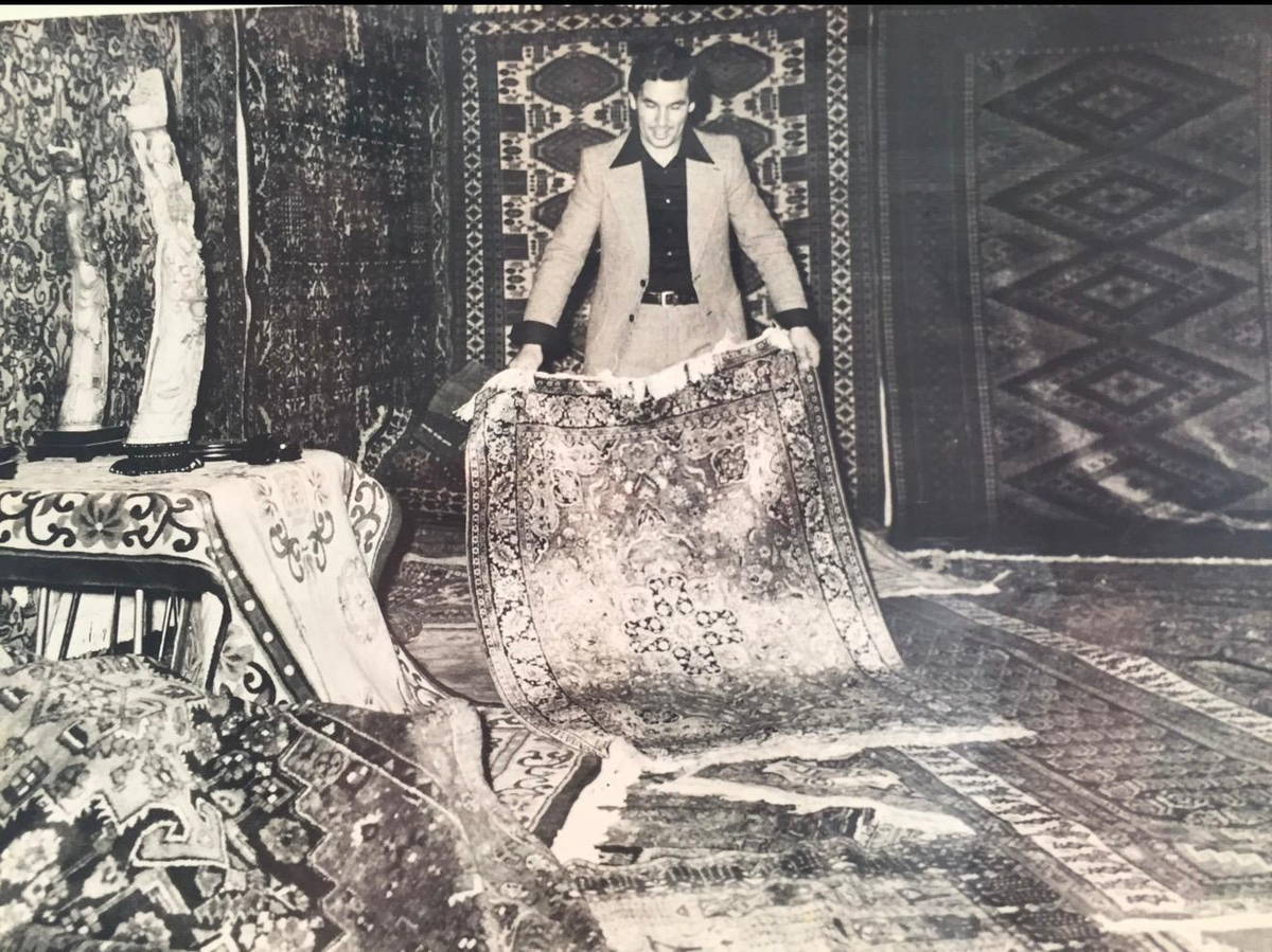 Photographie ancienne : un marchand déploie un tapis persan dans la boutique familiale, entouré de pièces suspendues
